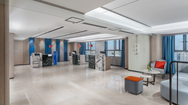 A modern hotel lobby with marble reception desks, a seating area with a sofa and ottoman, large windows with city views, and a high ceiling with recessed lighting.