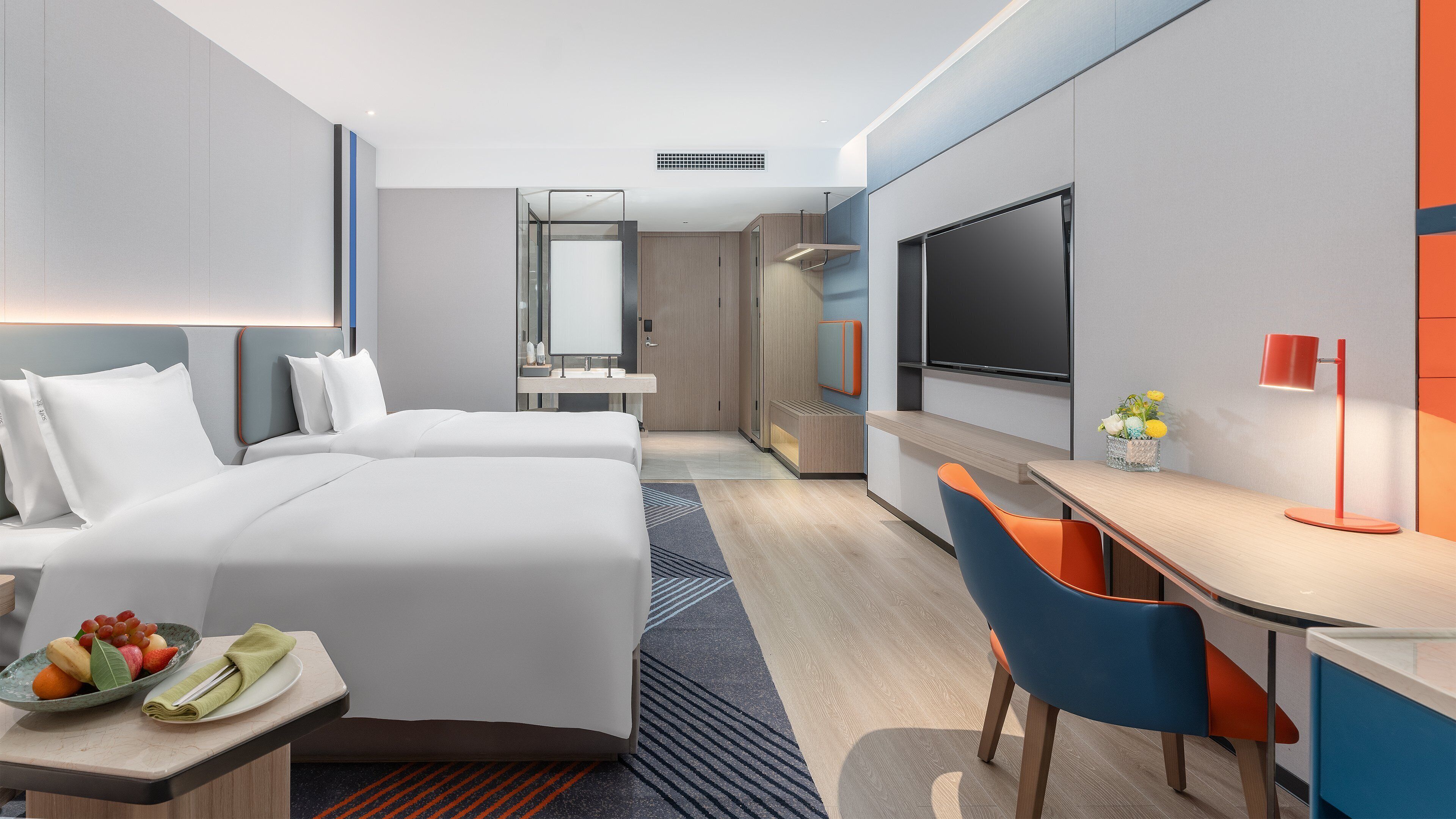 A hotel room with a bed, a desk, a chair, a TV, and a small table with fruit.