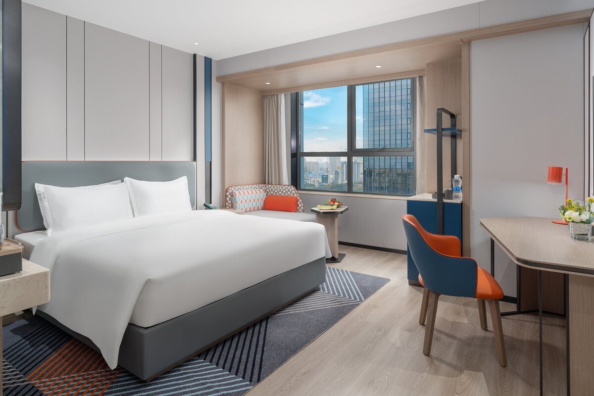Ein modernes Hotelzimmer mit einem großen Bett, einem Schreibtisch, einem Sessel, einem Fernseher und einem Fenster mit Blick auf die Stadt.