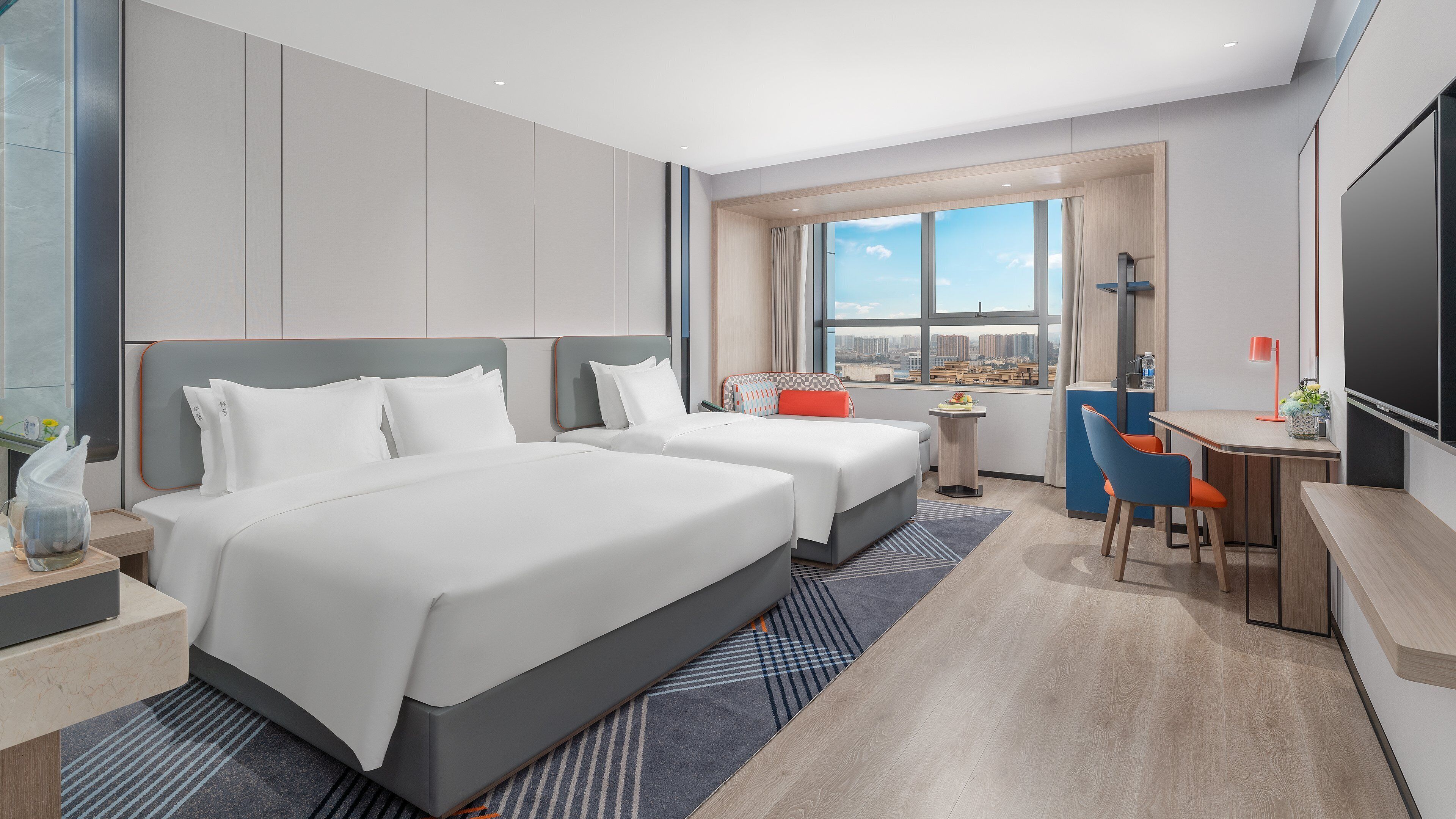 Habitación de hotel con dos camas, un escritorio, una silla, un televisor y un ventanal con vistas a la ciudad.