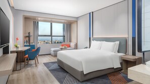 A modern hotel room with a large bed, a desk with a chair, a small table with a lamp, and a view of the city through large windows.