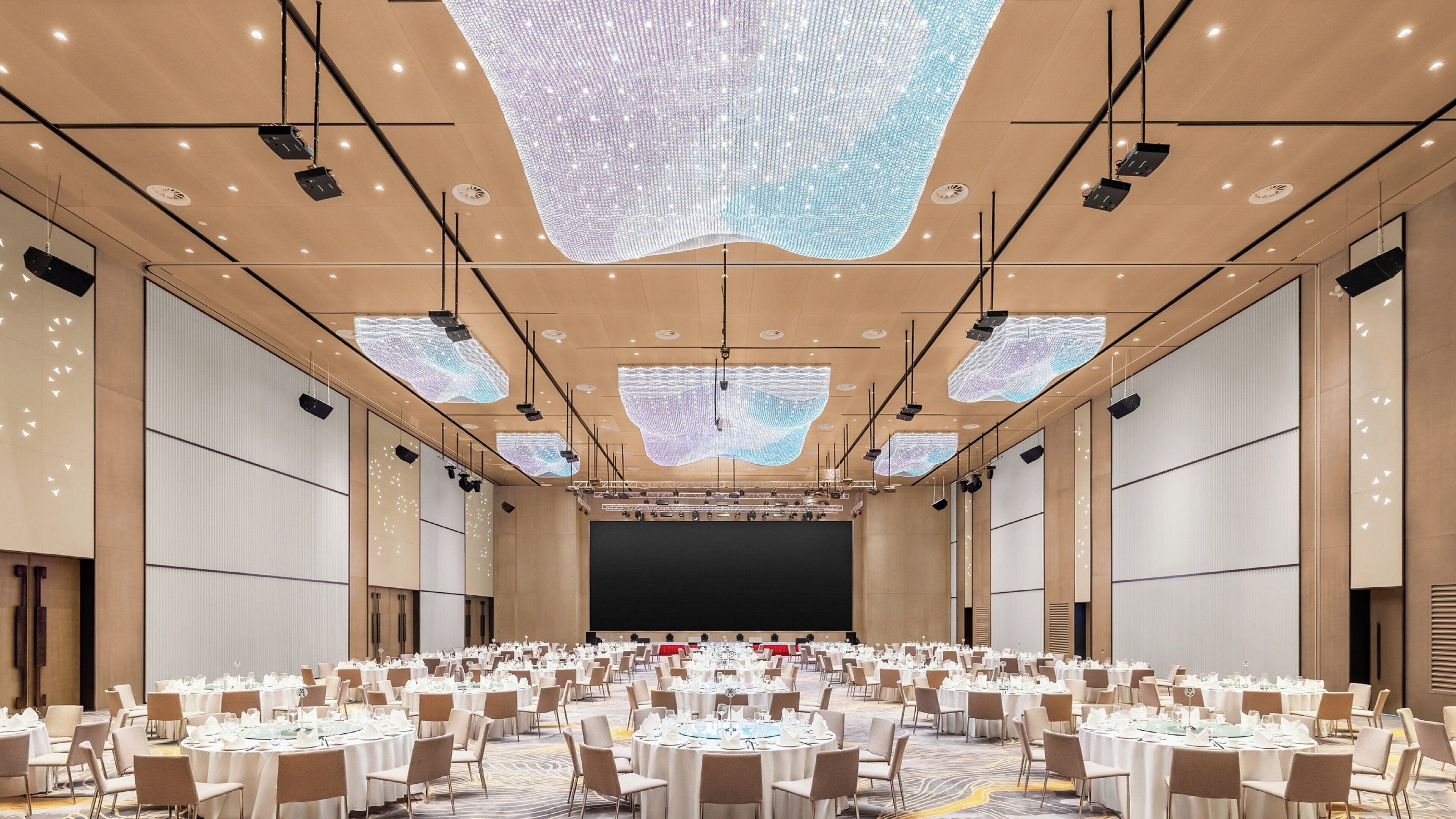 Un amplio salón de conferencias con mesas redondas dispuestas para comidas, equipado con proyectores y una pantalla grande al frente.