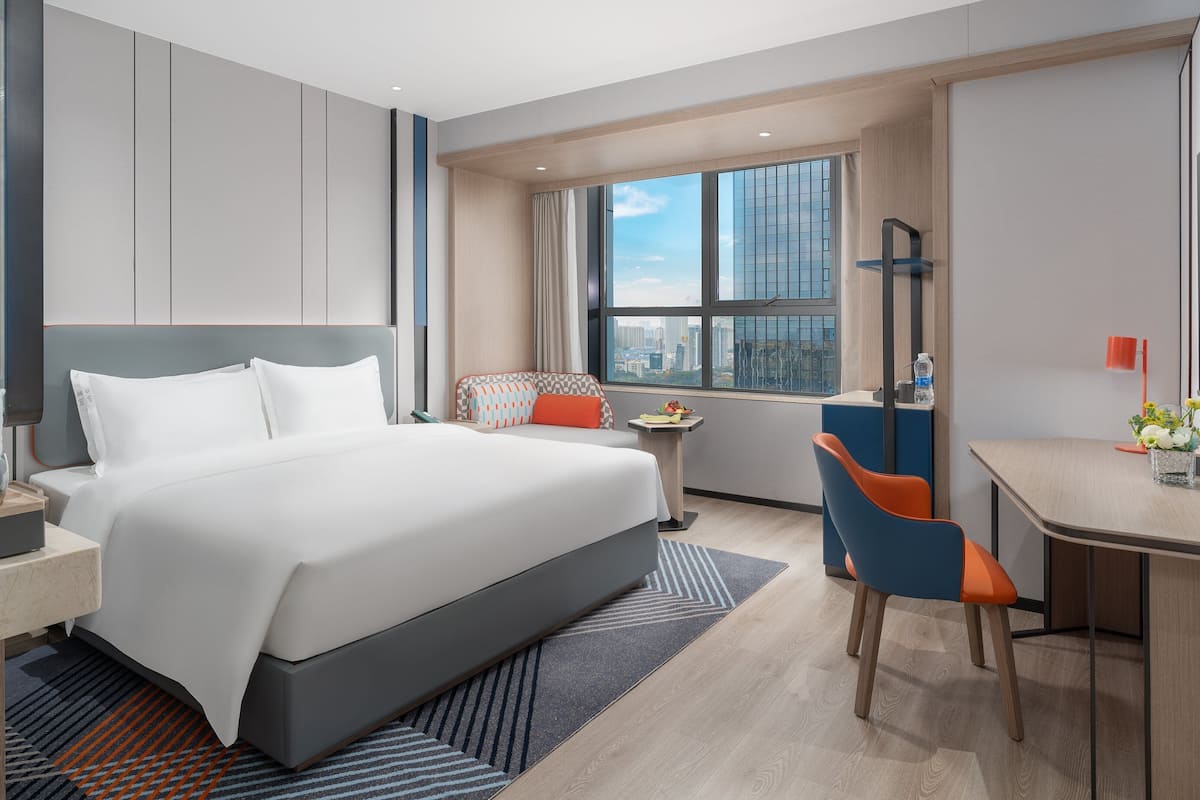 A modern hotel room with a large bed, a desk, a chair, a TV, and a window with a city view.