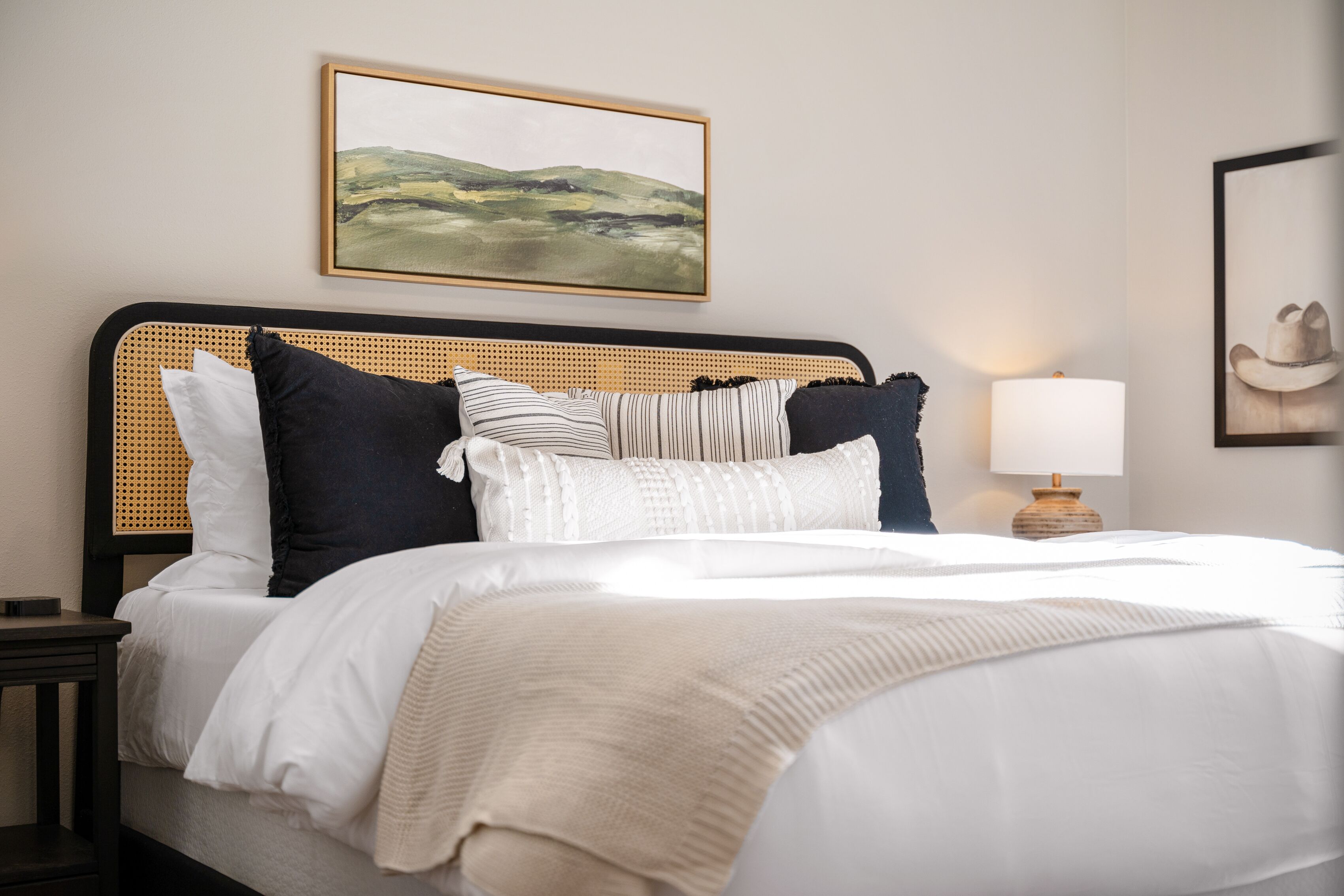 Una cama bien hecha con un cojín a rayas, uno blanco y uno negro. Hay una lámpara en una mesita de noche y un cuadro en la pared.