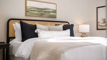 A neatly made bed with a striped pillow, a white pillow, and a black pillow. There is a lamp on a bedside table and a framed picture on the wall.