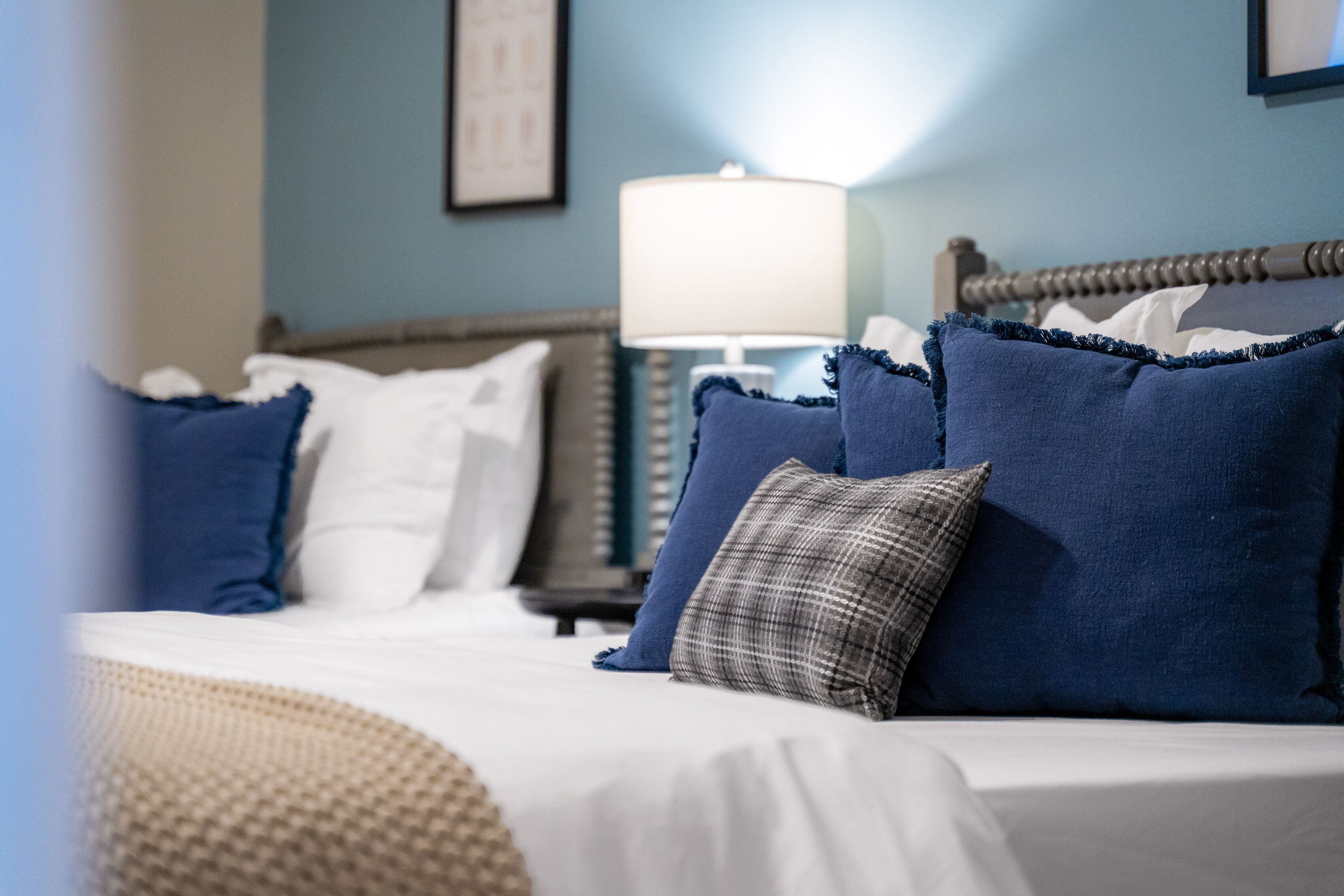 Ein ordentlich bezogenes Bett mit blau-weißen Kissen, einer Nachttischlampe und einem gerahmten Bild an der Wand.