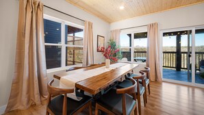 A dining area with a wooden table and chairs, large windows, and a view of the outdoors.