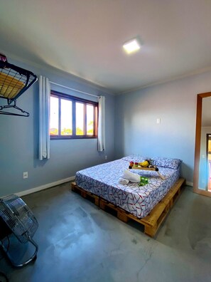 A bedroom with a bed on a wooden platform, a window with curtains, a ceiling fan, and a radiator. - RiqueHouse Hidro Piscina a 500m da praia (Marica)