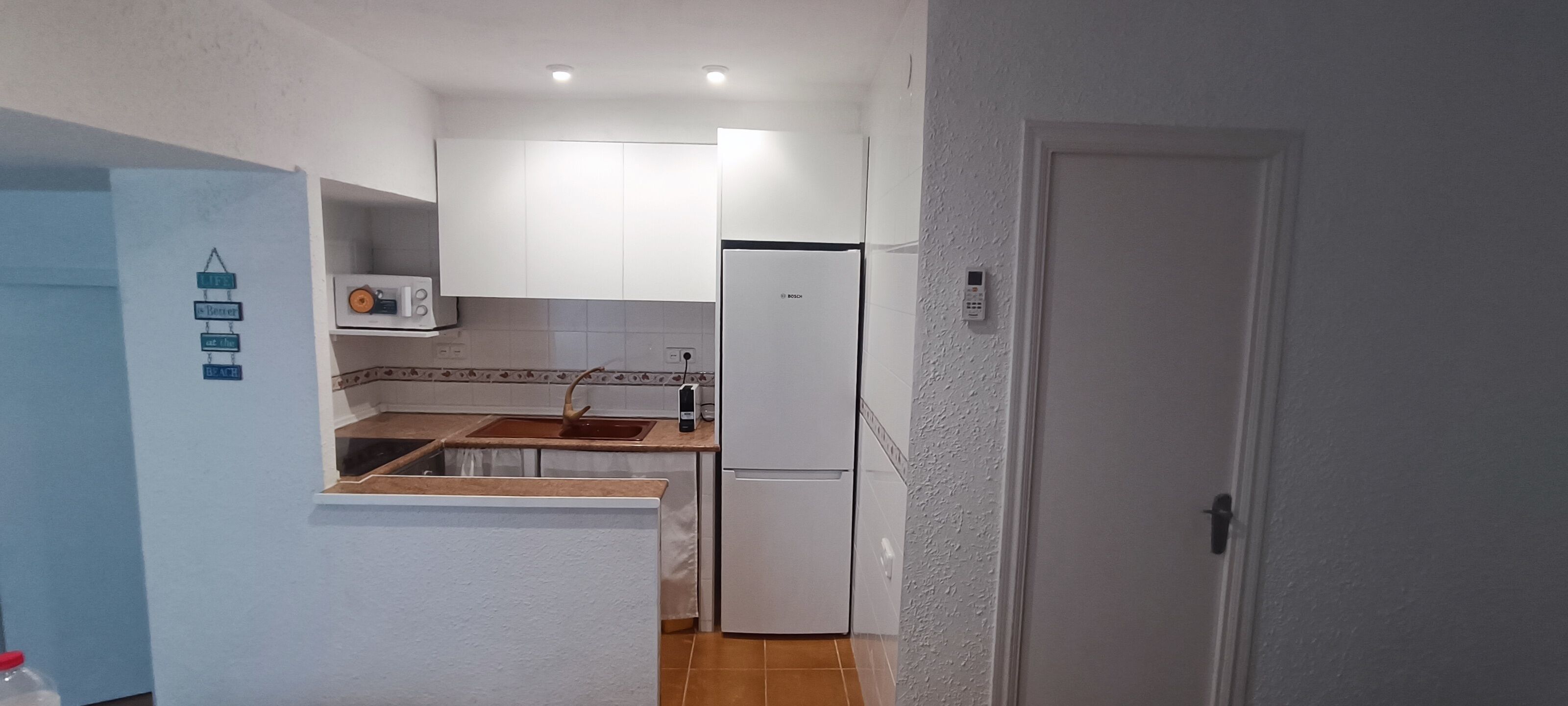 Een kleine keuken met witte kastjes, een magnetron, een spoelbak en een koelkast.