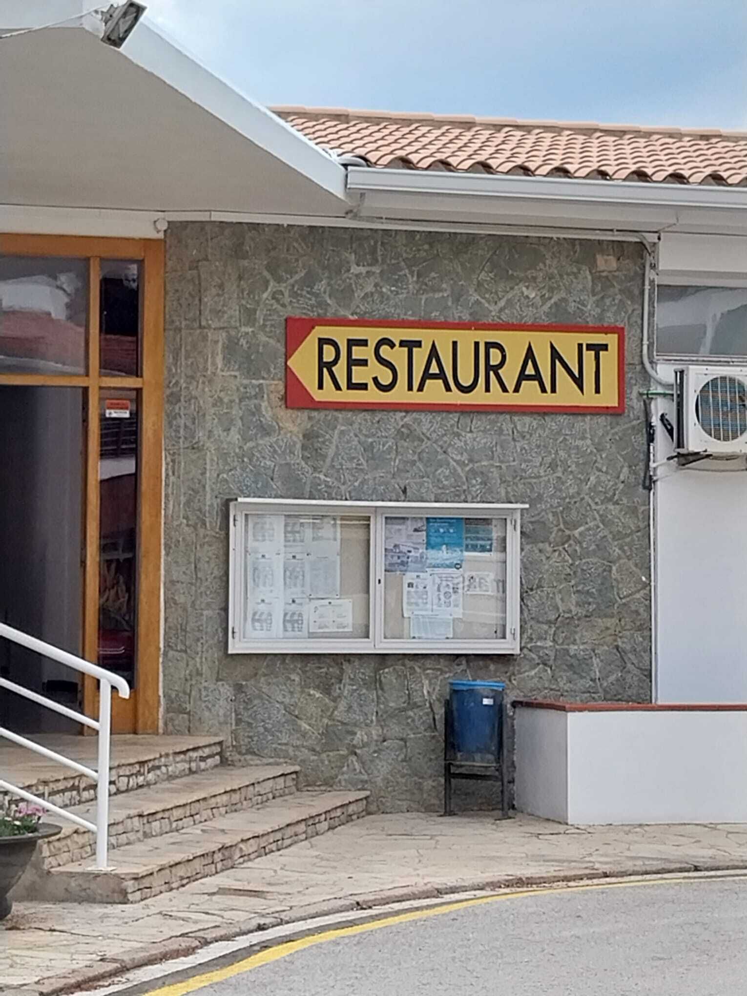 Un bâtiment à la façade en pierre, une enseigne indiquant un restaurant, une fenêtre avec des papiers et une poubelle bleue.