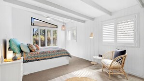 Une chambre à coucher avec un grand lit, un fauteuil en osier et une vue par une fenêtre équipée de stores.
