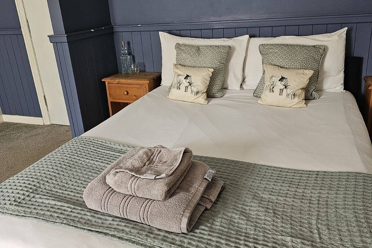 Una cama bien hecha con una toalla doblada, dos almohadas estampadas y una mesita de noche con un recipiente de vidrio.