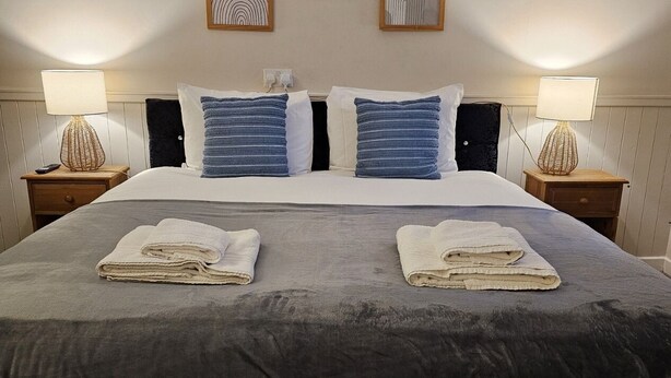 A neatly made bed with two folded towels, a dark grey bedspread, and a white pillow. There are two bedside lamps with decorative bases, a wall-mounted artwork above the bed, and a wooden nightstand with a remote control. - Dog and Partridge Inn (Calne)