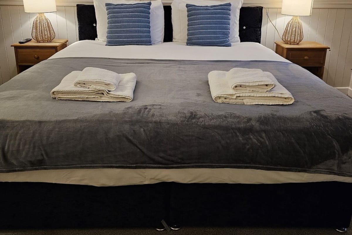 Una cama bien hecha con dos toallas dobladas, una colcha gris oscuro y una almohada blanca. Hay dos lámparas de noche con bases decorativas, una obra de arte en la pared sobre la cama y una mesita de noche de madera con un control remoto.