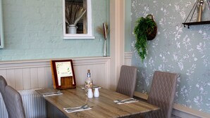 A dining area with a wooden table, four upholstered chairs, a menu frame, salt and pepper shakers, a bottle, and a hanging plant. - Dog and Partridge Inn (Calne)