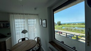 A dining area with a round table and chairs, a window with a view of a lake and greenery, and a ceiling light. - Bunk 21 Heiligenhafen Studio-Apartment (Heiligenhafen)