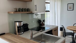 A modern kitchen with a built-in stove, a coffee machine, and a sink. There are wooden shelves above the counter and a dining area with a table and chairs. - Bunk 21 Heiligenhafen Studio-Apartment (Heiligenhafen)