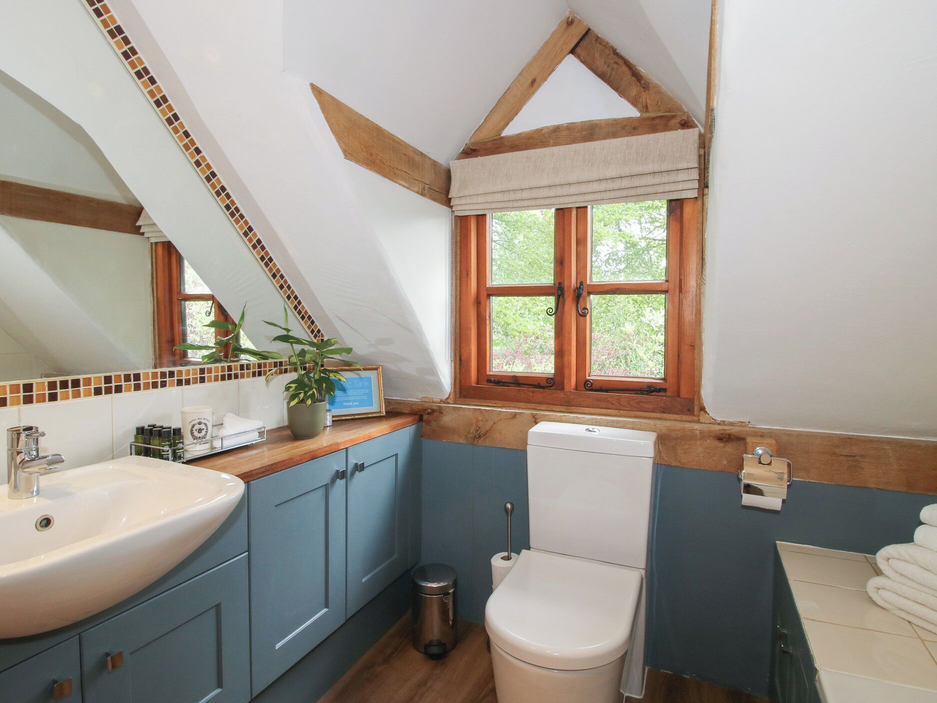 Een badkamer met een wit wastafelmeubel, blauwe kastjes, een wit toilet, een raam met uitzicht op groen en een houten plafond.