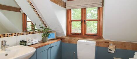Een badkamer met een wit wastafelmeubel, blauwe kastjes, een wit toilet, een raam met uitzicht op groen en een houten plafond.