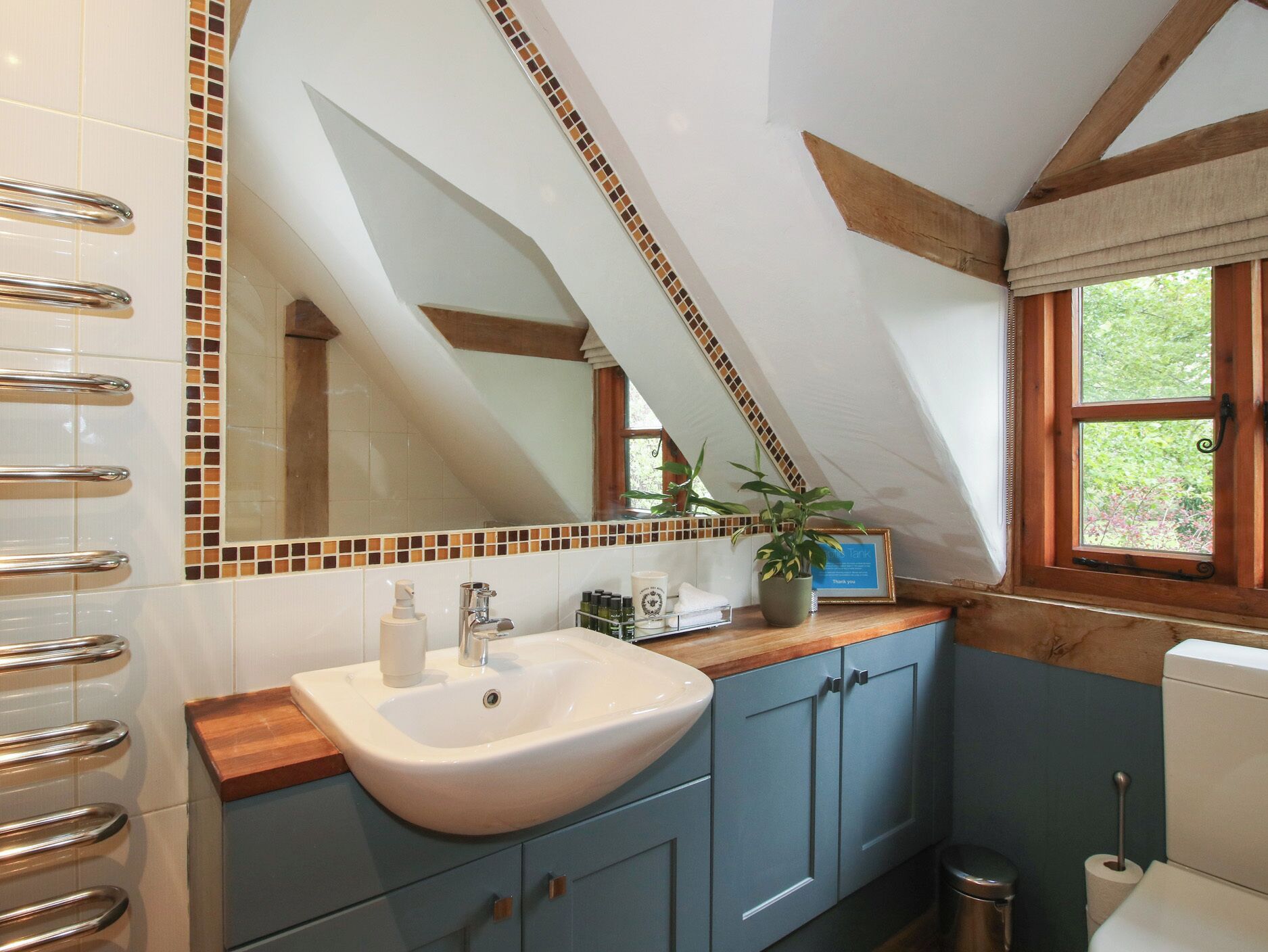 Un baño con lavamanos blanco, armarios azules, un espejo con detalles de mosaicos y una ventana con vistas a la vegetación.