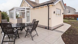 Una casa con un invernadero, un patio con mesa y sillas de vidrio, y un jardín.