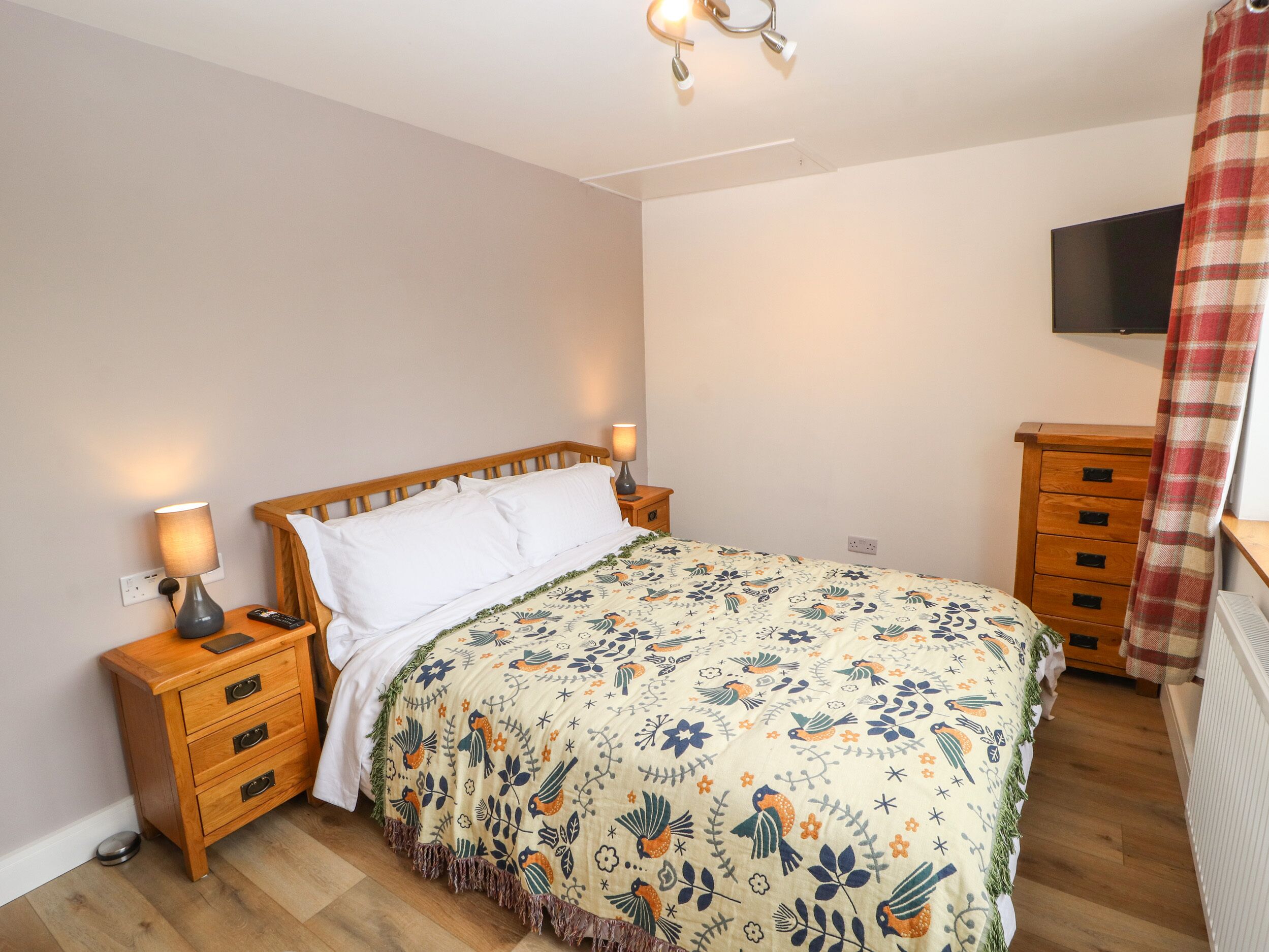 A bedroom with a wooden bed, two bedside tables with lamps, a TV on the wall, and a plaid curtain.