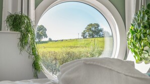 A room with a round window, a bed, and a view of a green field with trees. - Step into indulgence, where every detail is tailored for your comfort. (Cradley, near Malvern)