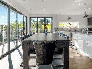A modern kitchen with a large island, a dining area with a long table and chairs, and large windows offering a view of the outdoors. - Step into indulgence, where every detail is tailored for your comfort. (Cradley, near Malvern)