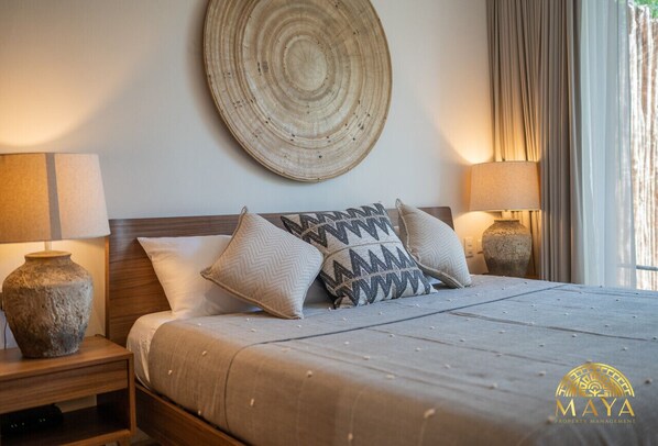 Uma cama bem arrumada com almofada estampada, duas luminárias de cabeceira, um enfeite de parede em tear e um vaso sobre a mesa de cabeceira.