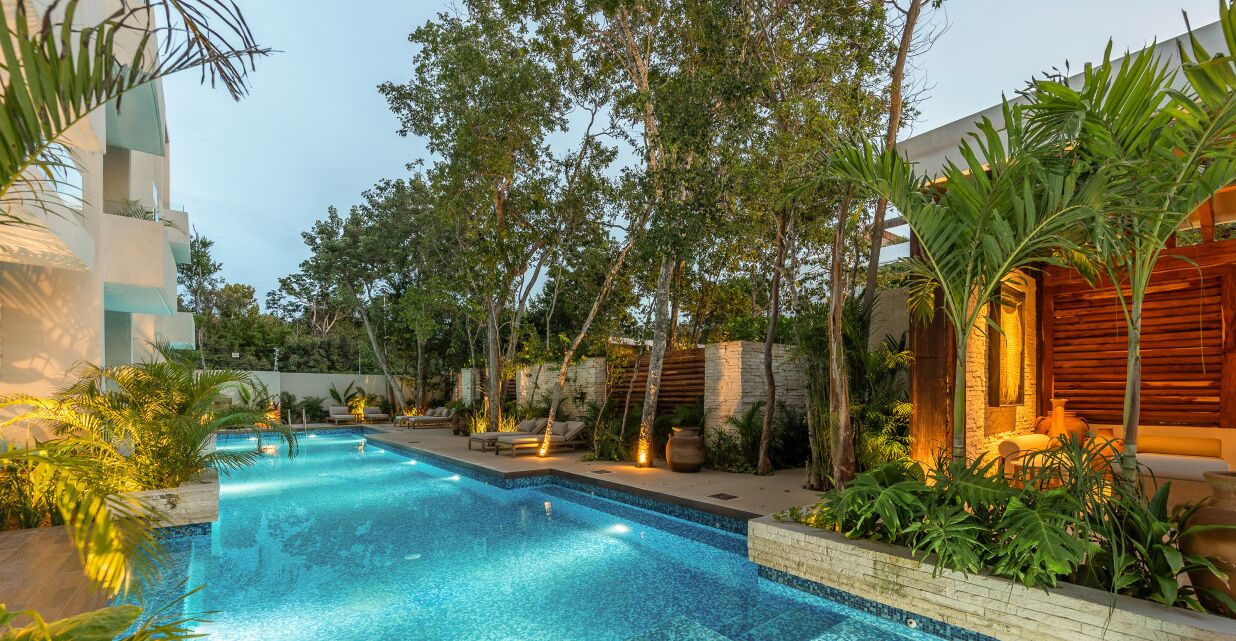 A swimming pool with lounge chairs and a wooden pergola, surrounded by lush greenery and a stone wall.