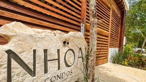 A stone sign with "NHOA CONDOS" written on it, placed in front of a wooden building with slatted walls. - NHOA 108C Lux 1BR Condo Plunge Pool in Aldea Zama (Tulum)