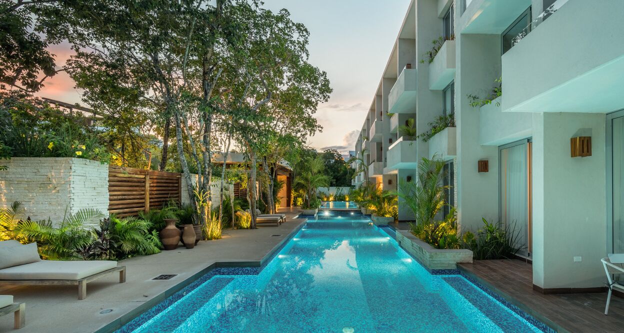 A swimming pool with lounge chairs and greenery, surrounded by a building with balconies and a stone wall.