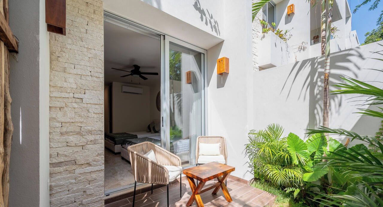 A patio with two wicker chairs and a wooden table, a sliding glass door leading to a bedroom, and a small garden area with tropical plants.