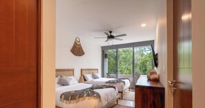 A hotel room with two beds, a ceiling fan, a large window, and a view of greenery outside.