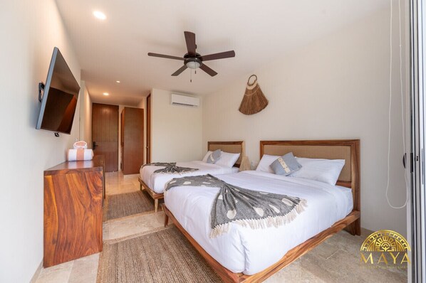 A hotel room with two beds, a wooden dresser, a ceiling fan, a TV mounted on the wall, and a woven basket on the wall. - NHOA 208C Lux 1BR Condo Jungle View in Aldea Zama (Tulum)
