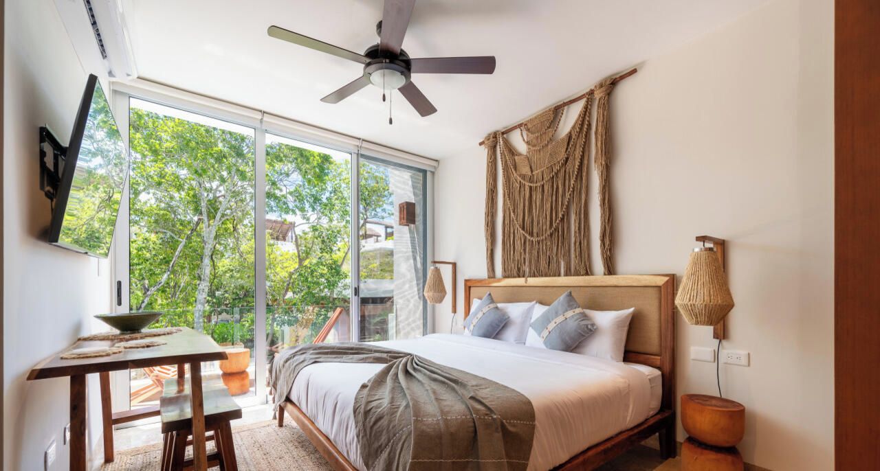 Un dormitorio con cama, escritorio, ventilador de techo y un amplio ventanal.
