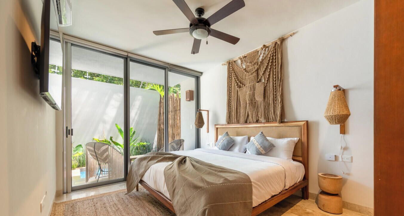 Un dormitorio con cama, ventilador de techo, ventana grande y vista a zonas verdes.