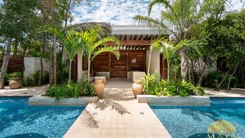Un área de piscina con una pérgola de madera, plantas tropicales y un edificio blanco de fachada de madera.