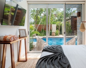 A bedroom with a large bed, a TV mounted on the wall, a small table with towels, and a view of an outdoor swimming pool.