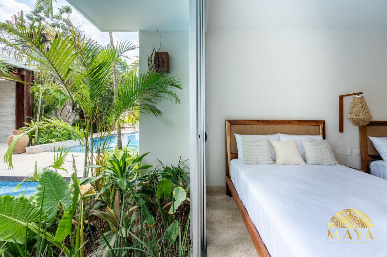 A bedroom with a wooden bed, white bedding, and a view of a swimming pool with tropical plants.