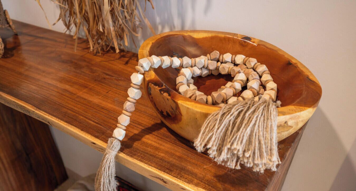 A wooden bowl with a beaded necklace and a tassel on a wooden table.