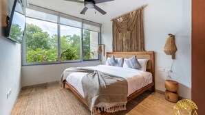 A bedroom with a bed, a ceiling fan, a large window with a view of greenery, a wall-mounted TV, and a macrame wall hanging.