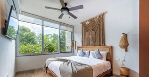 Une chambre avec un lit, un ventilateur de plafond, une grande fenêtre donnant sur un paysage verdoyant, un téléviseur fixé au mur et une décoration murale en macramé.
