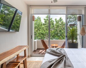 Een moderne hotelkamer met een groot bed, een houten bankje, een klein tafeltje en een balkon met uitzicht op bomen.