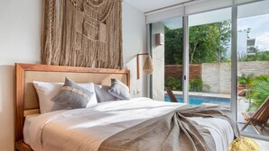 A bedroom with a large bed, a macrame wall hanging, and a view of a swimming pool and greenery outside. - NHOA 101B Lux 1BR Condo Pool access in Aldea Zama (Tulum)