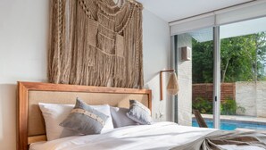A bedroom with a large bed, a macrame wall hanging, and a view of a swimming pool and greenery outside. - NHOA 101B Lux 1BR Condo Pool access in Aldea Zama (Tulum)