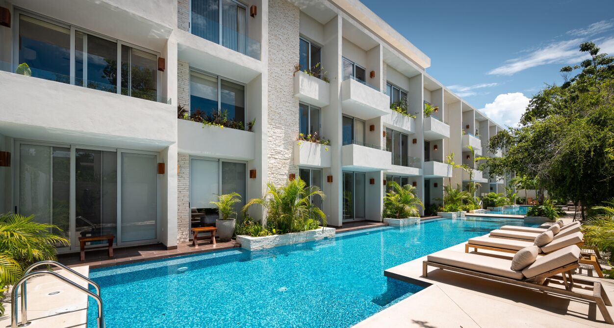 A modern hotel with a large outdoor pool, multiple balconies with plants, and a clear blue sky.
