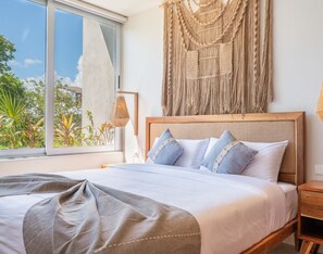 Een slaapkamer met een groot bed, een macramé wanddecoratie, een wandlamp en uitzicht op groen buiten.