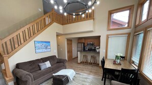 Ein geräumiger Wohnbereich mit einem braunen Sofa, einem Hocker und einem Esstisch mit Stühlen. Eine Treppe mit Holzwangen führt auf eine erhöhte Ebene.