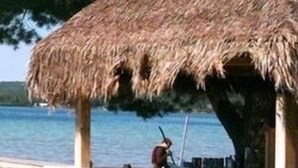 Eine Bar im Freien mit einem Reetdach, Holzhockern und Blick auf das Wasser.